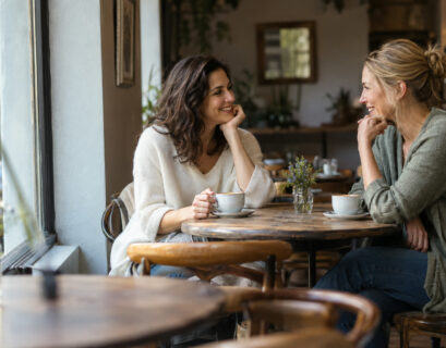 Two women laughing over coffee at a sunlit cafe table by a window